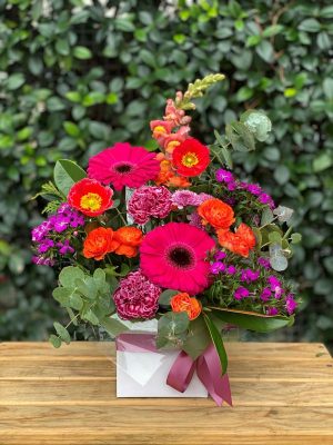 Boxed flower arrangement on wooden table and leafy background, red, purple and orange flowers and greenery, white box with purple ribbon bow